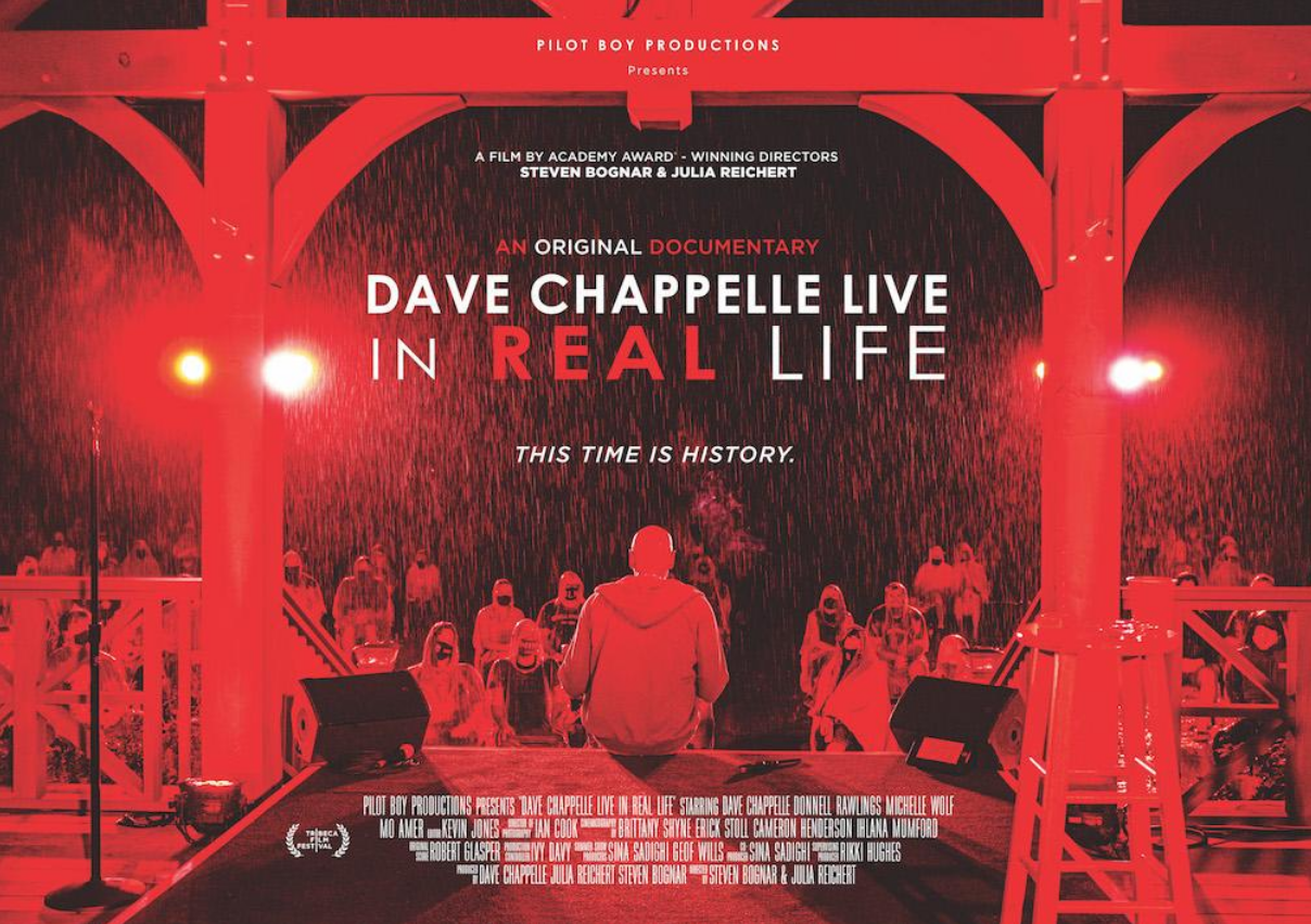film poster of dave chappelle live in real life. red hue we look out from backstage and see dave leaning against the stage and the audience faces. it is raining